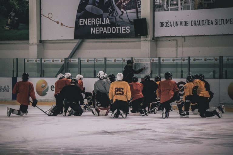 Eishockeytrainer gibt Anweisungen auf einer Taktiktafel am Spielfeld, die Mannschaft kniet im Halbkreis auf der Eisfläche