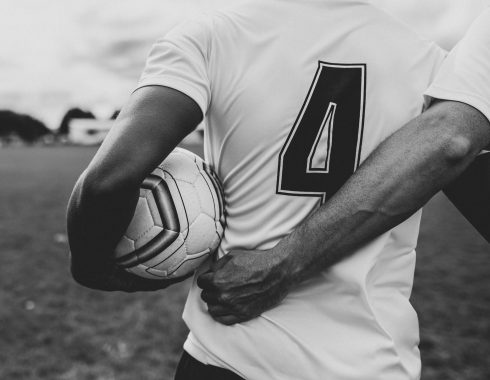 schwarzweißes Foto von Fußballer Rückenansicht, der den Ball in der linken Hand trägt und sich rechts mit be einem Teamkollegen einhängt